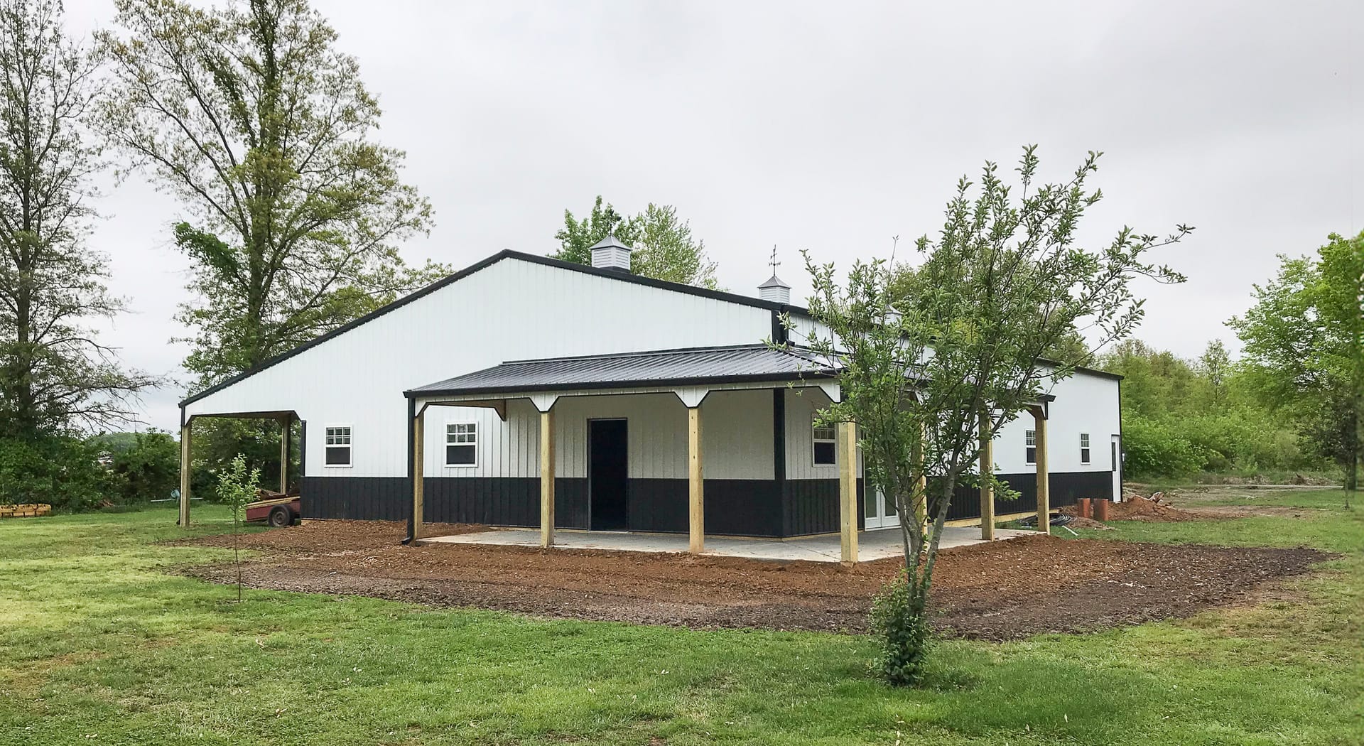 The image depicts a modern post-frame building set in a grassy area. The structure features a gable roof with two cupolas and a covered porch area supported by wooden posts. The building has a clean, two-tone exterior, with white upper walls and black low